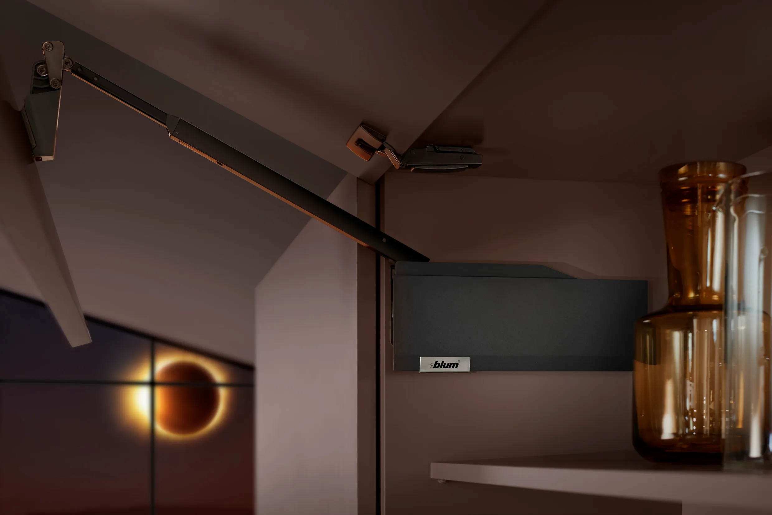 Detailaufnahme eines schwarzen Klappenbeschlags mit Hebelarm im geöffneten Oberschrank, dahinter Gläser in Bernsteinton