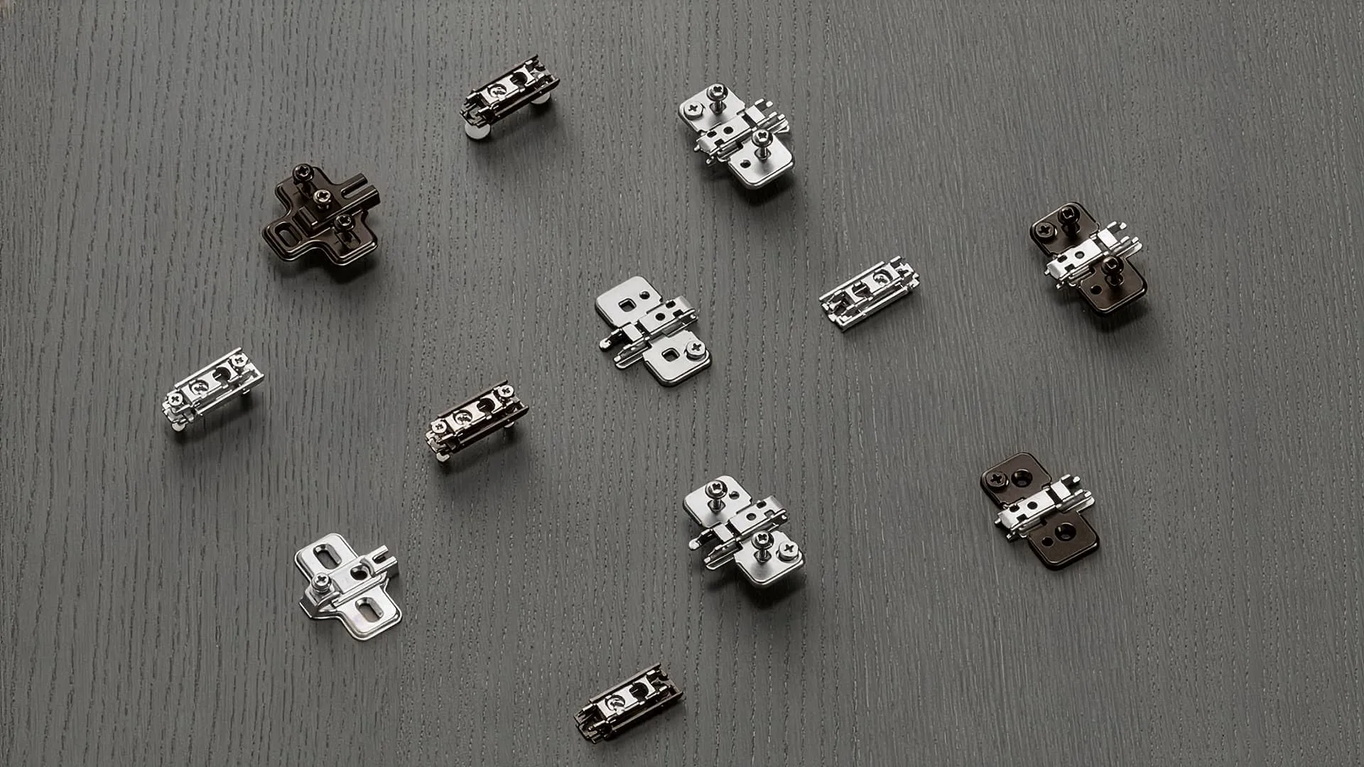 Verschiedene Blum-Montageplatten in Kreuz- und Linearform, Nickel und Onyx black, auf dunkler Holzfläche