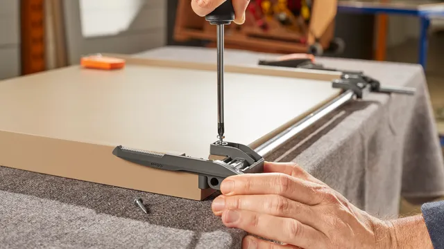 Hand mit Schraubendreher montiert eine Blum-Verriegelungseinheit auf der Unterseite eines Tablars, daneben die Synchronisationswelle.