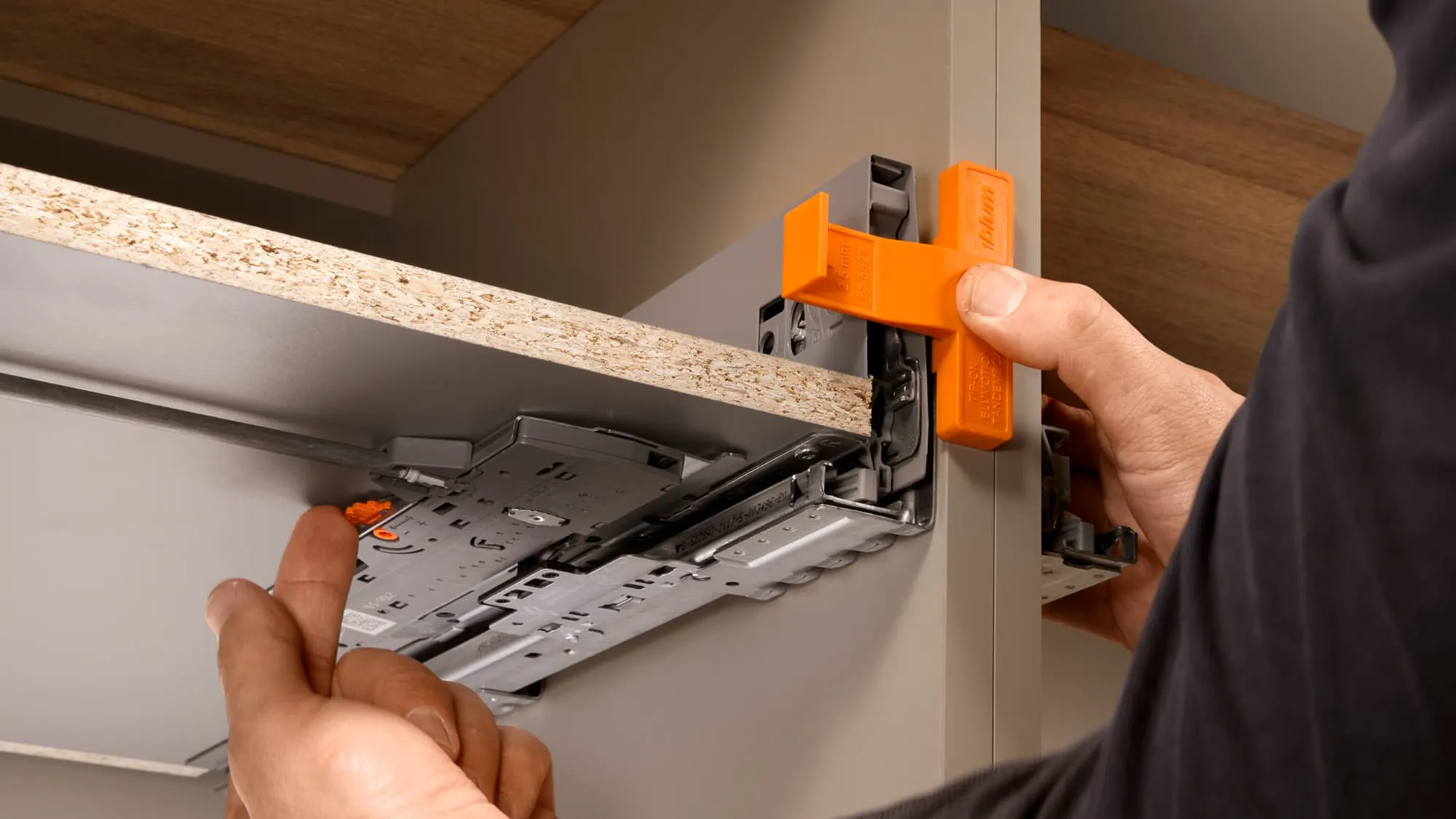 Zwei Hände stellen mit der orangen Blum-Einstelllehre das TIP-ON BLUMOTION an der Unterseite eines Schubkastens ein